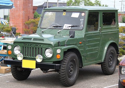 Dark green boxy small 4x4 with a steel roof, round headlamps, a slotted grille and auxiliary lamps, on a car park by a brick building. Opens a larger view