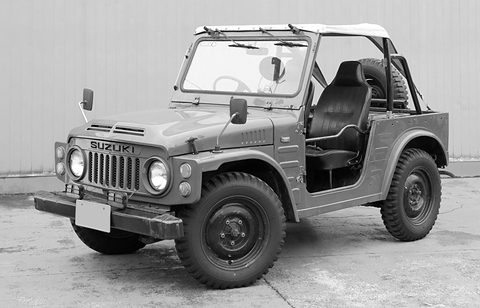 Black and white picture of a small open 4x4 with a soft-top frame, round headlamps and a slotted grille, against a corrugated wall. Opens a larger view