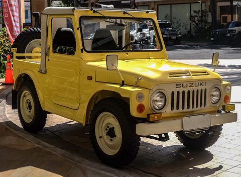 Pale yellow small 4x4 pick-up with a soft-top cab, round headlamps, a slotted grille and cream wheels, parked on paving. Opens a larger view
