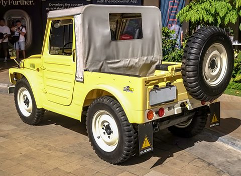 Rear quarter of a pale yellow small 4x4 pick-up with a grey canvas top, a side-mounted spare wheel and mud flaps, on paving. Opens a larger view