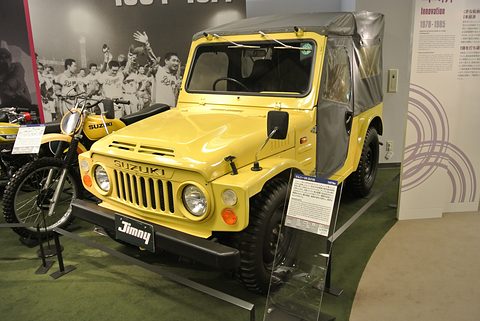 Yellow small 4x4 with a soft top and a Jimny plate on its bumper, on a museum floor with motorcycles and a placard beside it. Opens a larger view