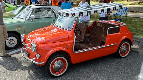 Coral-red doorless Fiat 500 with brown wicker seats, a striped fringed awning and red-rimmed whitewall wheels. Opens a larger view