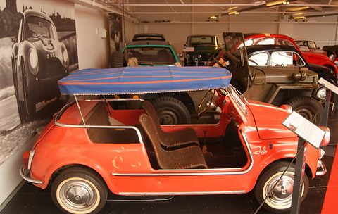 Orange doorless Fiat 500 with two wicker seats and a blue fabric awning, in a museum hall with a large period photograph behind. Opens a larger view