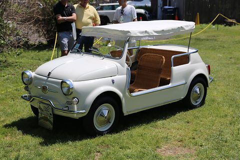 White doorless Fiat 500 with wicker seats, a white fringed awning and raised round headlamps, parked on grass.