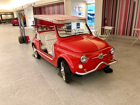 A red doorless Fiat 500 beach car with white seats and a fringed pink-striped canopy in a hotel lounge. Opens a larger view