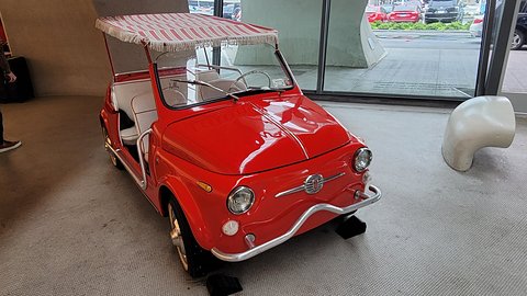 Red doorless Fiat 500 with a pink and white striped fringed awning and white seats, beside a glass wall. Opens a larger view