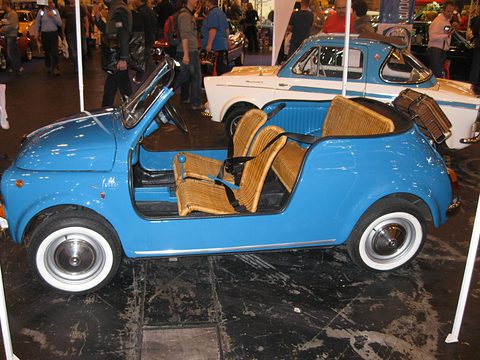 Side view of a light blue doorless Fiat 500 with cane seats and a wicker hamper on the tail, crowds behind. Opens a larger view