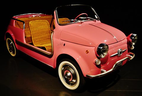 Pink doorless Fiat 500 with golden wicker seats and whitewall tyres, lit against a black background. Opens a larger view
