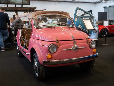 Pink doorless Fiat 500 with leather bonnet straps and a beige fringed awning, visitors at tables to its left. Opens a larger view