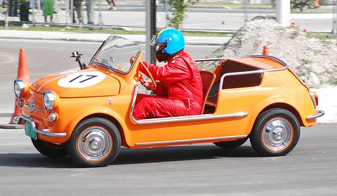 Orange doorless Fiat 500 with race number 17, its driver in a red suit and blue helmet, cornering past traffic cones. Opens a larger view
