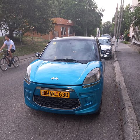 Head-on view of a bright turquoise small hatchback with an orange number plate on a tree-lined street. Opens a larger view