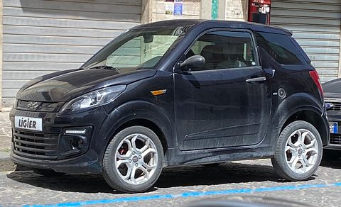 Dusty black small three-door hatchback with silver five-spoke wheels and a LIGIER grille plate, parked by a shuttered shop.