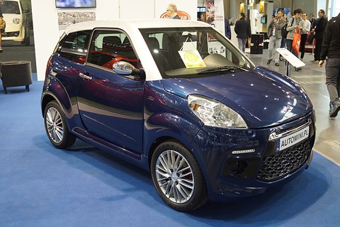 Dark blue three-door microcar with a white roof and silver alloy wheels on blue carpet, visitors walking behind.