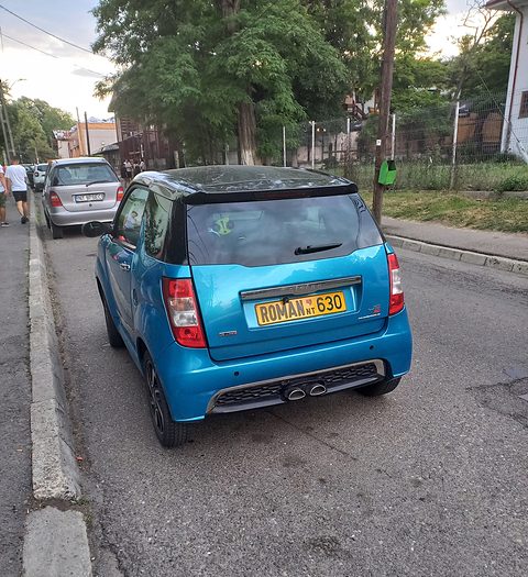 Rear view of a turquoise small hatchback with a black roof, twin tailpipes and an orange plate parked by a kerb. Opens a larger view