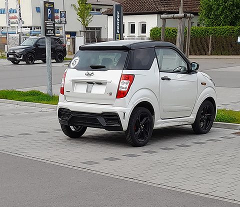Rear three-quarter view of a white small hatchback with a black roof and black wheels in a paved car park. Opens a larger view