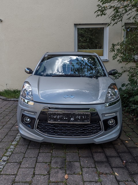 Head-on view of a silver small hatchback with swept headlamps and a black mesh grille in front of a cream house wall. Opens a larger view