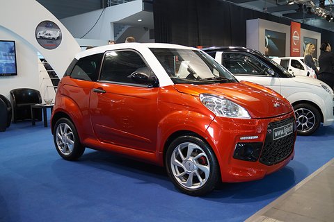 Orange small three-door hatchback with a white roof, swept-back headlamps and alloy wheels on blue show carpet.