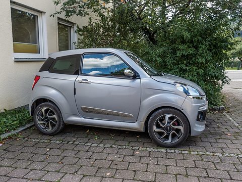 Side view of a short silver three-door car with dark alloy wheels parked on paving in front of a bush. Opens a larger view