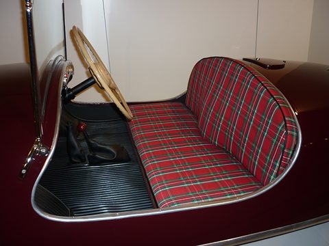 Inside a maroon open car: a red tartan bench seat, a large wood-rimmed steering wheel and a ribbed rubber floor. Opens a larger view