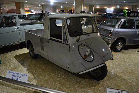 Grey three-wheeled pickup on a beige floor among parked cars under rows of ceiling lights. Opens a larger view