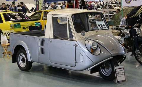 Front three-quarter of a grey three-wheeled pickup with a cream cab, its single front wheel under a rounded nose. Opens a larger view
