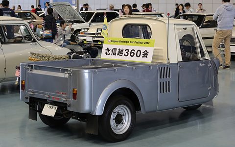 Rear three-quarter of a grey three-wheeled pickup with a cream cab and a club sign on its roof, in a busy hall. Opens a larger view
