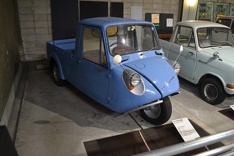 Blue three-wheeled pickup with a pale cab roof in a concrete museum bay, a white small car beside it. Opens a larger view