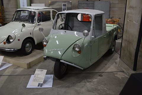Pale green three-wheeled pickup with a white cab on a museum floor, a white Subaru beside it. Opens a larger view