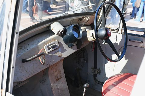 Inside a small truck cab: a black thin-rimmed wheel, a cream instrument pod, a lever and a red seat. Opens a larger view