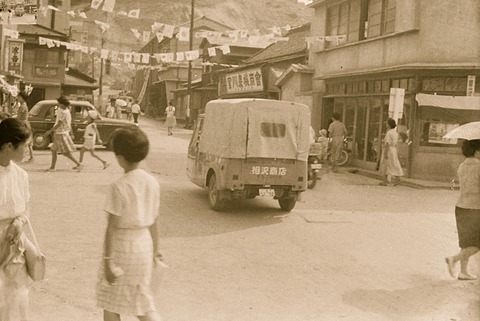 A sepia street scene: a small covered three-wheeled truck from behind among shoppers, flags strung overhead.