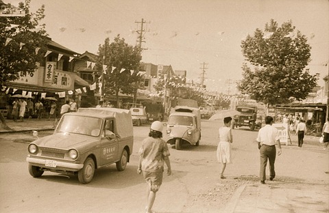 A sepia street scene with bunting overhead: a small pick-up in front, a three-wheeled truck behind it among shoppers. Opens a larger view