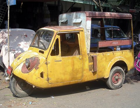 Battered yellow three-wheeled truck with flaking paint and a tall wooden box body, parked on a dusty street.