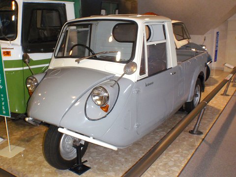 Grey three-wheeled pickup with a white cab on a speckled museum floor, a green bus at left. Opens a larger view
