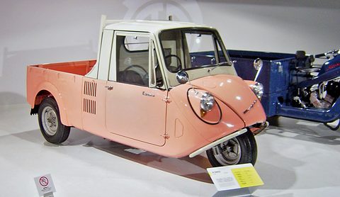 Salmon-orange three-wheeled pickup with a cream cab and one front wheel, a blue three-wheeler behind. Opens a larger view