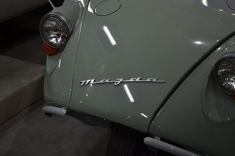 Close view of a pale green rounded nose with a chrome Mazda script between two round headlamps. Opens a larger view
