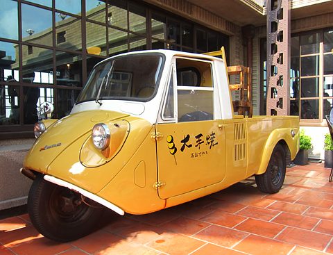 Yellow three-wheeled pickup with a white cab roof and black Chinese lettering on the door, on terracotta tiles. Opens a larger view