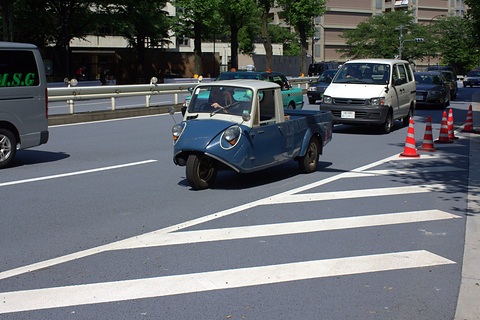 Small blue three-wheeled truck crossing a wide road with white markings, a white van and taxis behind. Opens a larger view