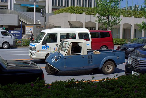Side view of a blue three-wheeled pickup with a cream cab beside a hedge, a white van behind. Opens a larger view