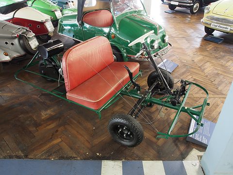 Green tubular frame on three small wheels carrying a red bench seat, steering column and engine.