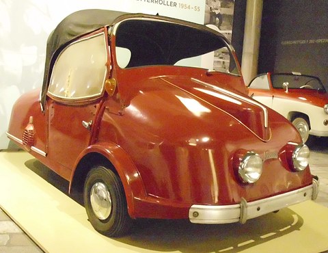 Maroon three-wheeled scooter-car with a black hood and twin headlamps on a pale plinth. Opens a larger view