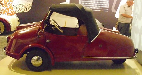 Side view of a maroon three-wheeler with a black fabric hood on a plinth, a visitor reading a display behind. Opens a larger view