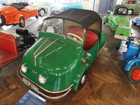 Green three-wheeler with black hood on a parquet floor among scooters and a Messerschmitt. Opens a larger view