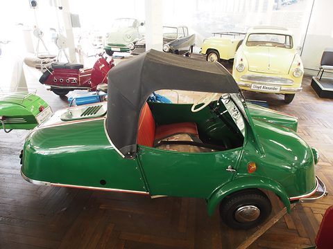 Green three-wheeler in side view with red seats and black hood, a pale yellow Lloyd behind. Opens a larger view
