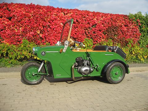 Green open three-wheeler with exposed single-cylinder engine on brick paving before red and green foliage. Opens a larger view