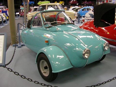Turquoise single-seat three-wheeler with a small windscreen behind a chain barrier among microcars.
