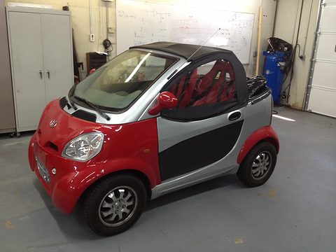 Red and silver two-seat microcar with a black soft top and open sides on a grey workshop floor. Opens a larger view