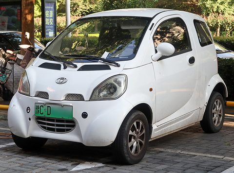 White two-door microcar with round headlamps and a green new-energy plate on paving in front of shrubs. Opens a larger view