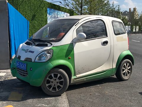 White two-door microcar with green lower panels and a blue Chinese plate parked by a blue hoarding.