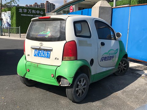 Rear of a white two-door microcar with green lower body and a K10 badge beside blue site hoardings. Opens a larger view