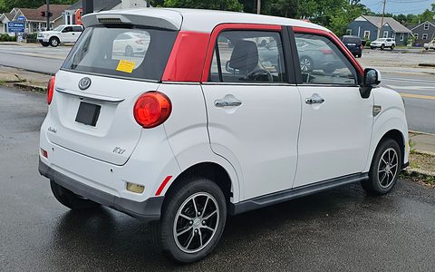 Rear of a white small four-door car with a red roof panel and round tail lamps on a wet car park. Opens a larger view
